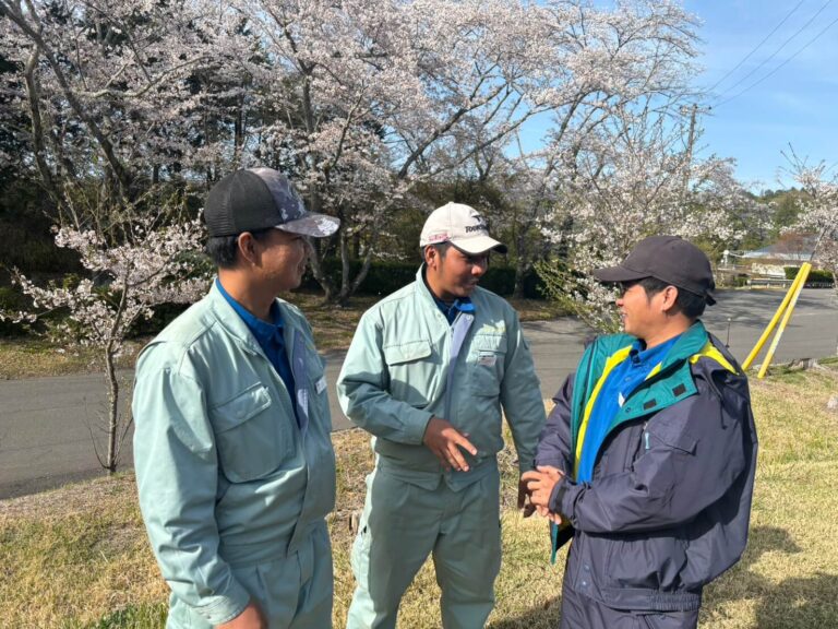 桜の咲くのゴルフ場で、作業服を着た3人の男性が屋外で会話をしている様子。