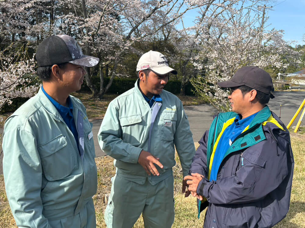 春の桜の下で談笑する作業服姿のフィリピン人スタッフたち。