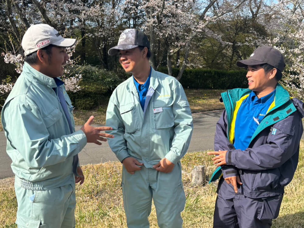 桜の木の下で話し合う作業服姿の3人のフィリピン人スタッフ。和やかな雰囲気。