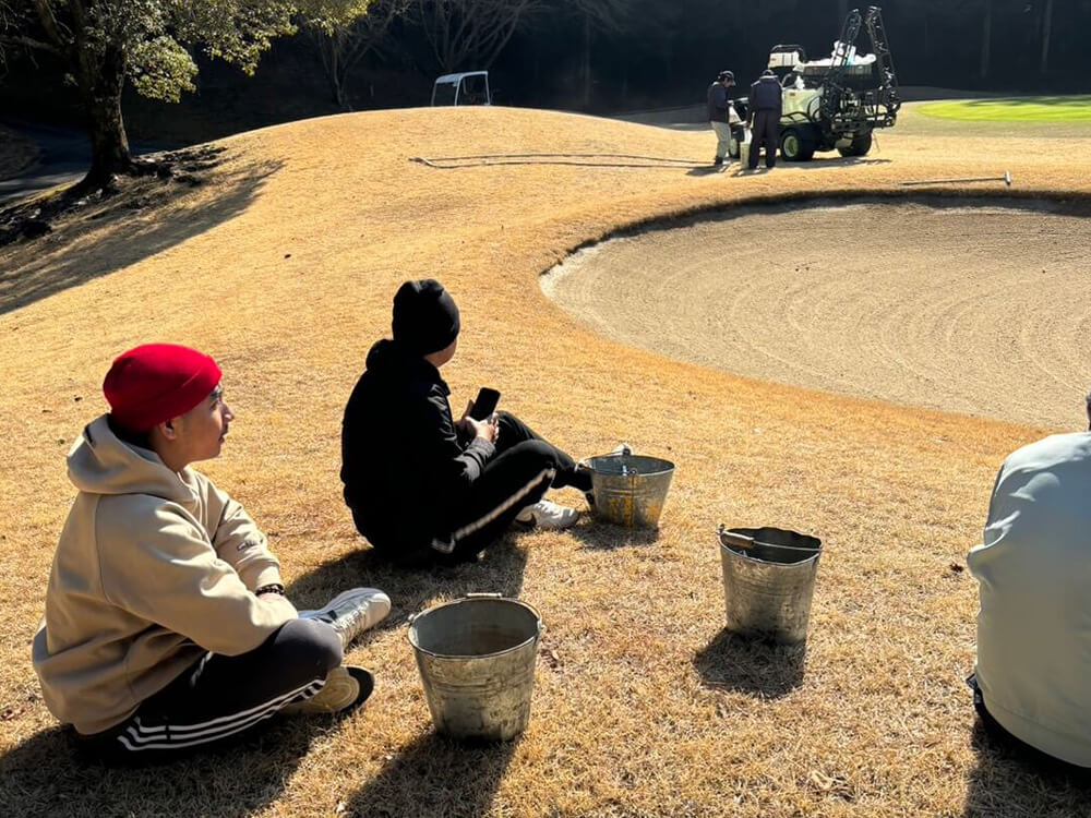 ルフ場で芝のメンテナンス作業を行う3人の作業員。地面にひざをついて真剣に作業している。